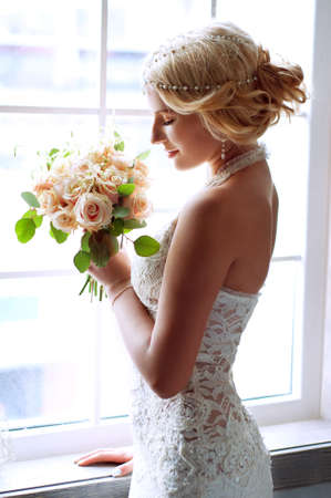 Charming blond bride waiting near the window for her groom wearing white lace dress and pearl...