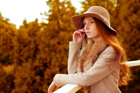 Woman with long red hair, fedora hat and fur coat standing on the balcony in the woods in autumn. Melancholic mood.の写真素材