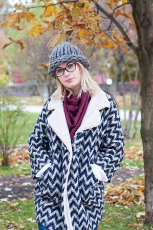 Student girl in autumn park wearing glasses, warm cardigan and thick knitted hat. Fall street style city look.の写真素材