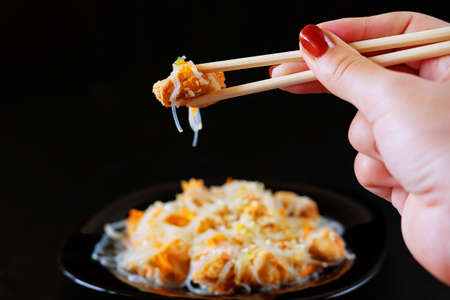 Asian traditional food. Fried rice noodles with chicken meat, vegetable slices sprinkled with sesame seeds.Hand holding a piece of meat in chopsticks.の写真素材