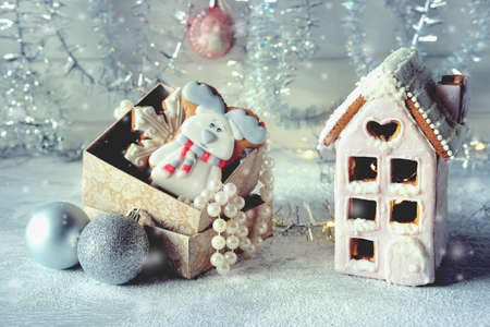 Magical winter christmas picture. Gingerbread house with pink icing with light shining in the windows under snow falling and new year toys and decorations in paper box with cookies.の写真素材