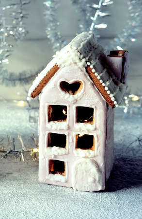Magical winter christmas picture. Gingerbread house with pink icing with light shining in the windows under snow falling and new year toys and decorations in paper box with cookies.の写真素材
