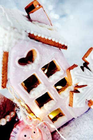 Magical winter christmas picture. Gingerbread house with pink icing with light shining in the windows under snow falling and new year toys and decorations in paper box with cookies.の写真素材