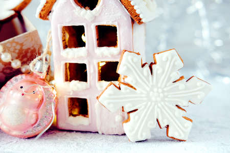 Magical winter christmas picture. Gingerbread house with pink icing with light shining in the windows under snow falling and new year toys and decorations in paper box with cookies.の写真素材