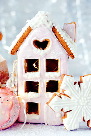 Magical winter christmas picture. Gingerbread house with pink icing with light shining in the windows under snow falling and new year toys and decorations in paper box with cookies.の写真素材