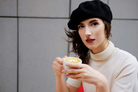 Stylish french girl in beret drinking coffee outdoors in the city. Casual portrait. の写真素材
