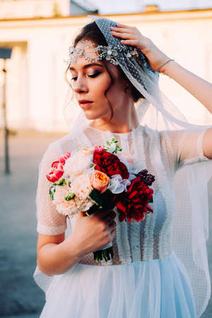 Beautiful eastern style bride with white veil and hairpiece holding bridal bouquet posing outdoors at sunsetの写真素材