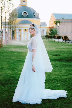 Charming young bride in white dress and veil walking to the church in the summer eveningの写真素材