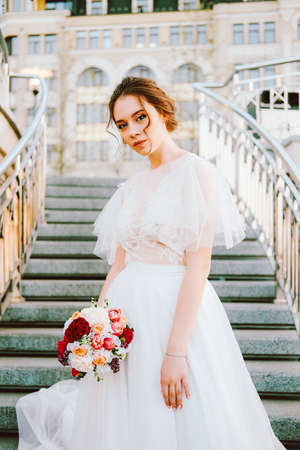Adorable caucasian bride in white flowing wedding dress made of lace and tulle standing on the staircase in sunset eveningの写真素材
