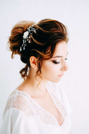 Tender elegant young brunette bride with hairdo, hairpin and bridal makeup wearing white peignoir....