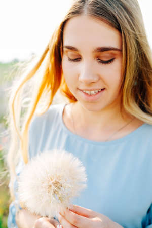 Young woman with long blond hair and blue dress holding dry dandelion flowerの写真素材