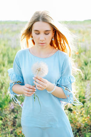 Young woman with long blond hair and blue dress holding dry dandelion flowerの写真素材