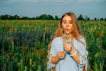 Young woman with long blond hair and blue dress holding dry dandelion flowerの写真素材
