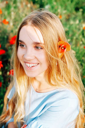 Calm happy blond woman with long straight hair and blue dress sitting in the field of poppy flowersの写真素材