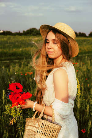 Happy young blond woman smiling in the field of poppies wearing linen jumpsuit, hat and woven basket with wind blown hairの写真素材