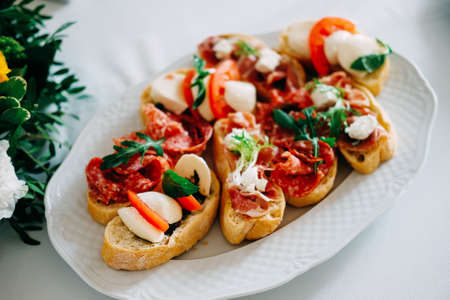 Italian bruschetta snack with ham, sausage, tomato, mozzarella, herbs served on white plate in restaurantの写真素材