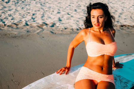 Young woman in white bikini at the sea shore resting at the old surf board