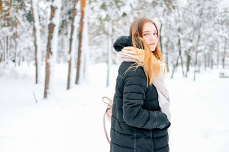Attractive blond young adult woman walking through winter forest full of snow wearing casual outfit of black parka, jeans, pink leather boots and backpack and beige shawlの写真素材