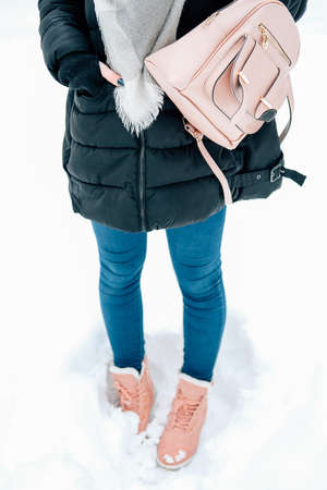 Attractive blond young adult woman walking through winter forest full of snow wearing casual outfit of black parka, jeans, pink leather boots and backpack and beige shawlの写真素材