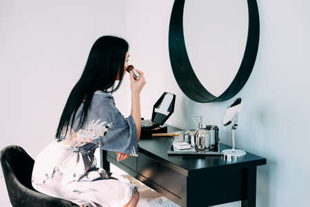 Beautiful brunette with long hair wearing bath robe, looking into the mirror and doing her makeupの写真素材
