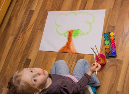 Overhead view of an attractive little girl kneeling on a hardwood floor painting a colourful tree on a sheet of paper with watercoloursの写真素材