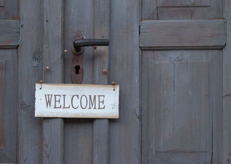 Welcome sign hanging below the handle on an old grey wooden door greeting guests as they enterの写真素材