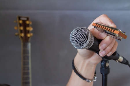 Close up of a hand holding a mouth organ and microphone during a musical performance or recital with a guitar visible in the background on a grey wallの写真素材