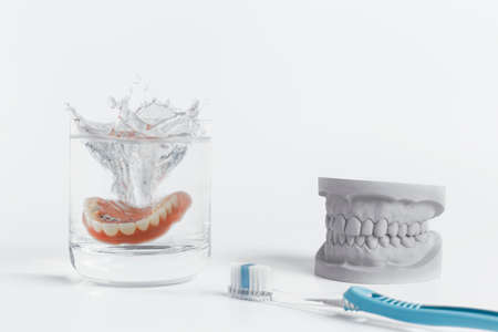 Dentures hygiene concept displaying mold falling into glass of water and pair of toothbrushes on white backgroundの写真素材