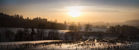 Panoramic view of a glowing orange sunrise over a cold snowy winter landscape with leafless deciduous trees on rolling hills and feint mist still clinging to the groundの写真素材