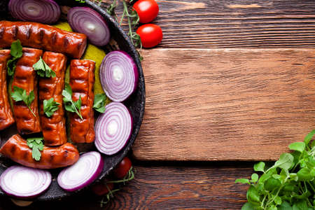 Grilled sausage in a pan, cutting board and fresh vegetables on wooden tableの写真素材