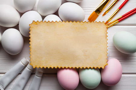 Blank paper sheet, colored and white eggs, brushes and paints on white wooden table の写真素材