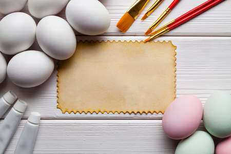 Blank paper sheet, colored and white eggs, brushes and paints on white wooden table の写真素材