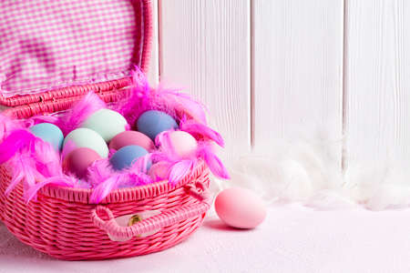 Happy Easter - pink basket full of colored eggs and feathers on white wooden backgroundの写真素材