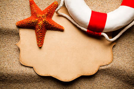 Orange starfish, red and white lifebuoy and blank paper label on beach sandの写真素材