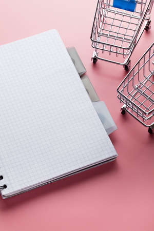 Two shopping carts and notebook with a blank page on pink tableの写真素材