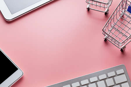 Shopping carts, computer keyboard and modern tablet on pink background