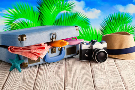 Suitcase full of holiday items - photo camera, summer hat, tanning oil, towel and flip flops on tropical backgroundの写真素材