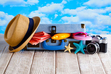 Open holiday suitcase; summer hat, towel, tanning oil, photo camera; flip flops on wooden tableの写真素材