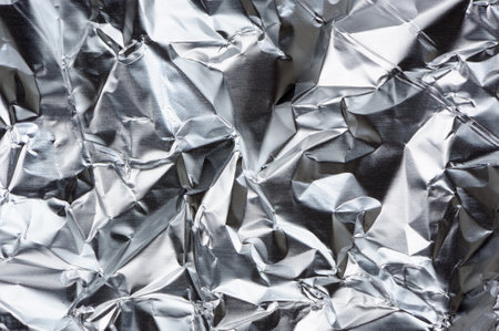A dynamic texture of crumpled aluminum foil forming a unique and reflective background, creating a captivating visual interplay of light and shadow with its metallic folds and creases.の写真素材