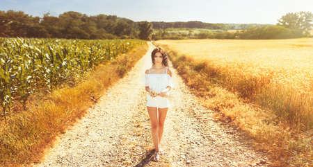 Beauty romantic girl enjoying nature in outdoors. Happy young woman in white shorts holding the ears on the road near of golden ripe wheat field in sun light. Free summer dreams.の写真素材