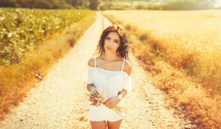 Beauty romantic girl enjoying nature in outdoors. Happy young woman in white shorts holding the ears on the road near of golden ripe wheat field in sun light. Free summer dreams.の写真素材
