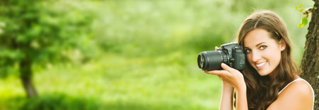 Beautiful smiling woman is professional photographer with DSLR camera and taking pictures outdoors in summer nature. Wide banner.の写真素材