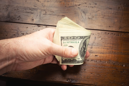 Hand holding one hundred US banknotes over wooden table background. Cash of hundred dollar bills,...