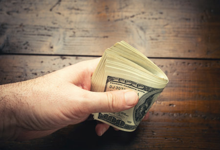 Hand holding one hundred US banknotes over wooden table background. Cash of hundred dollar bills, paper money currency.の写真素材