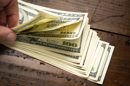 Hand counting pile of one hundred US banknotes on wooden table background. Cash of hundred dollar...