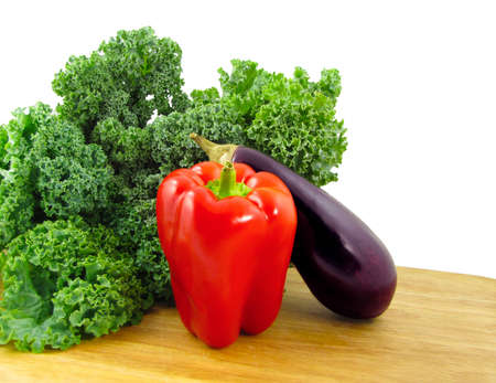 A red bell pepper, a purple eggplant, and a bunch of green kale on a wooden cutting board isolated...