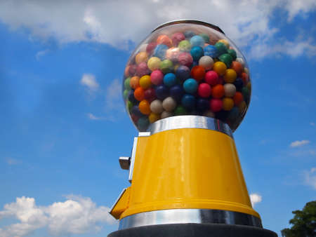 A vintage yellow gumball machine with multi-colored gumballs stands tall in front of a blue sky with wispy clouds.の写真素材