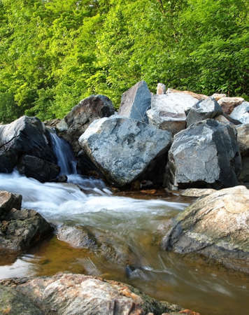 Small waterfalls rush through a woodland stream with large rocks and green foliage の写真素材