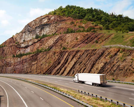 A large white trailer truck drives through a mountain pass with steep sedimentary rock to the sides of the road の写真素材