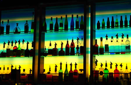 A multi-color glass wall behind a bar illuminates a collection of alcohol bottles of many interesting shapes in silhouette standing on glass shelves in dark, ambient bar at night.のeditorial素材